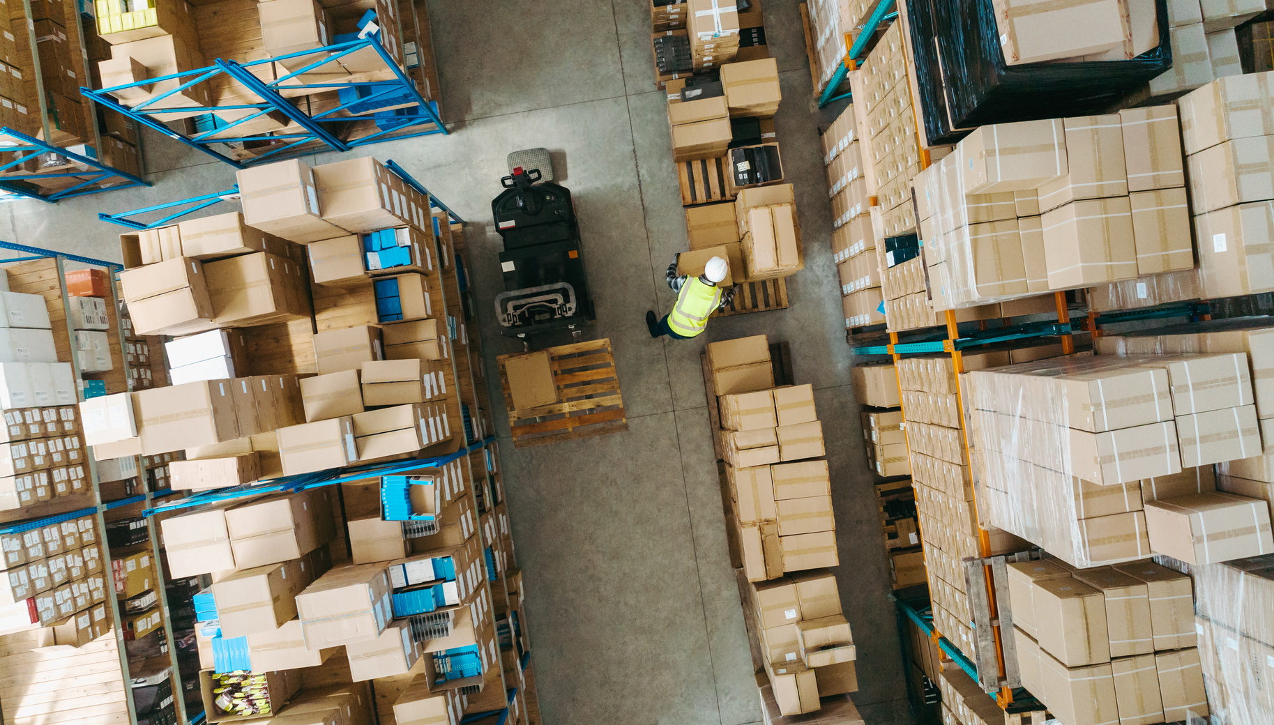 Aerial photo of a warehouse worker picking boxes. They are wearing a hi-vis vest and surrounded by boxes on both sides.