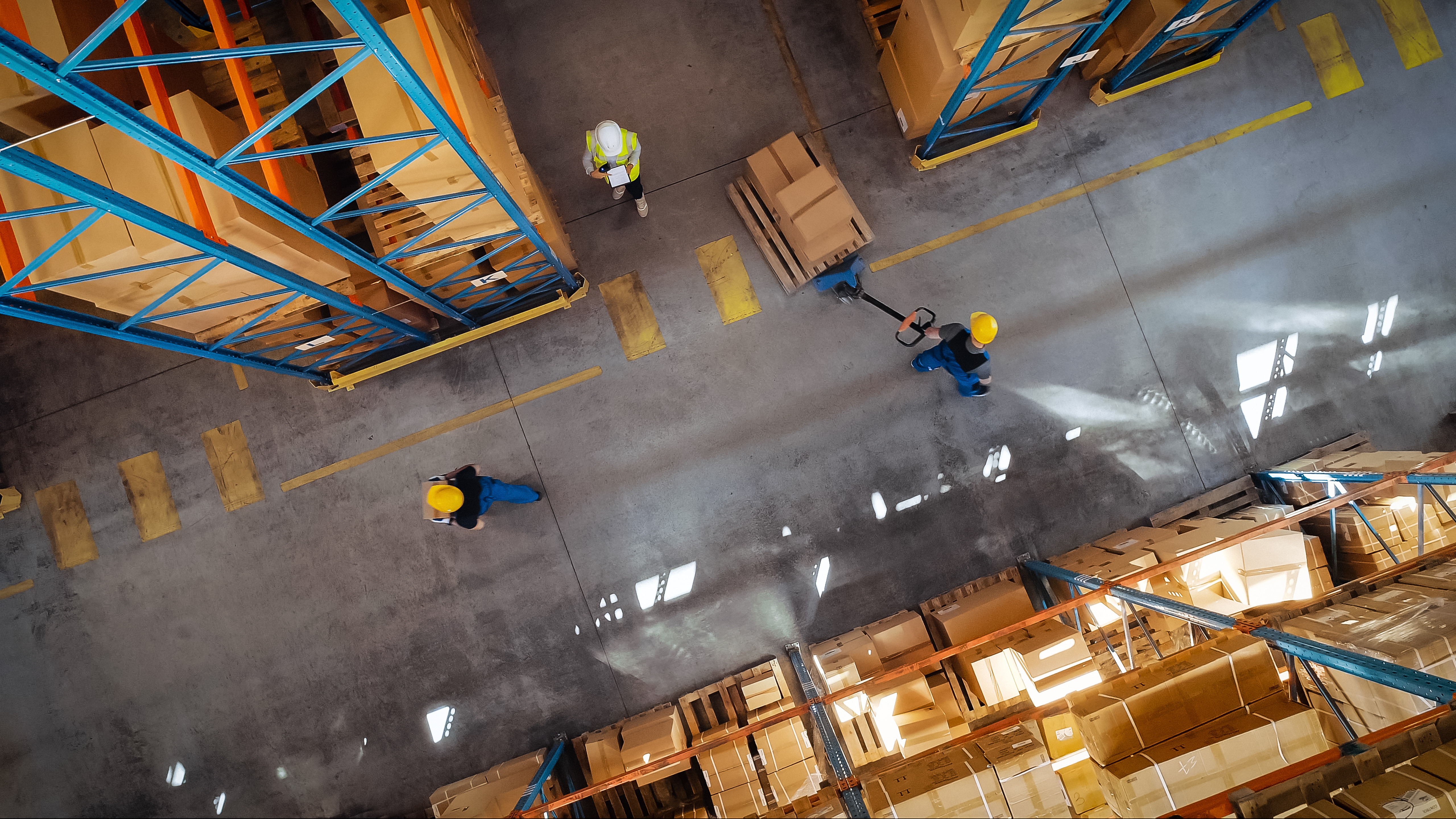 Aerial photo of a warehouse worker moving boxes on a trolley. They are wearing blue overalls and a yellow hard hat and surrounded by boxes on both sides.